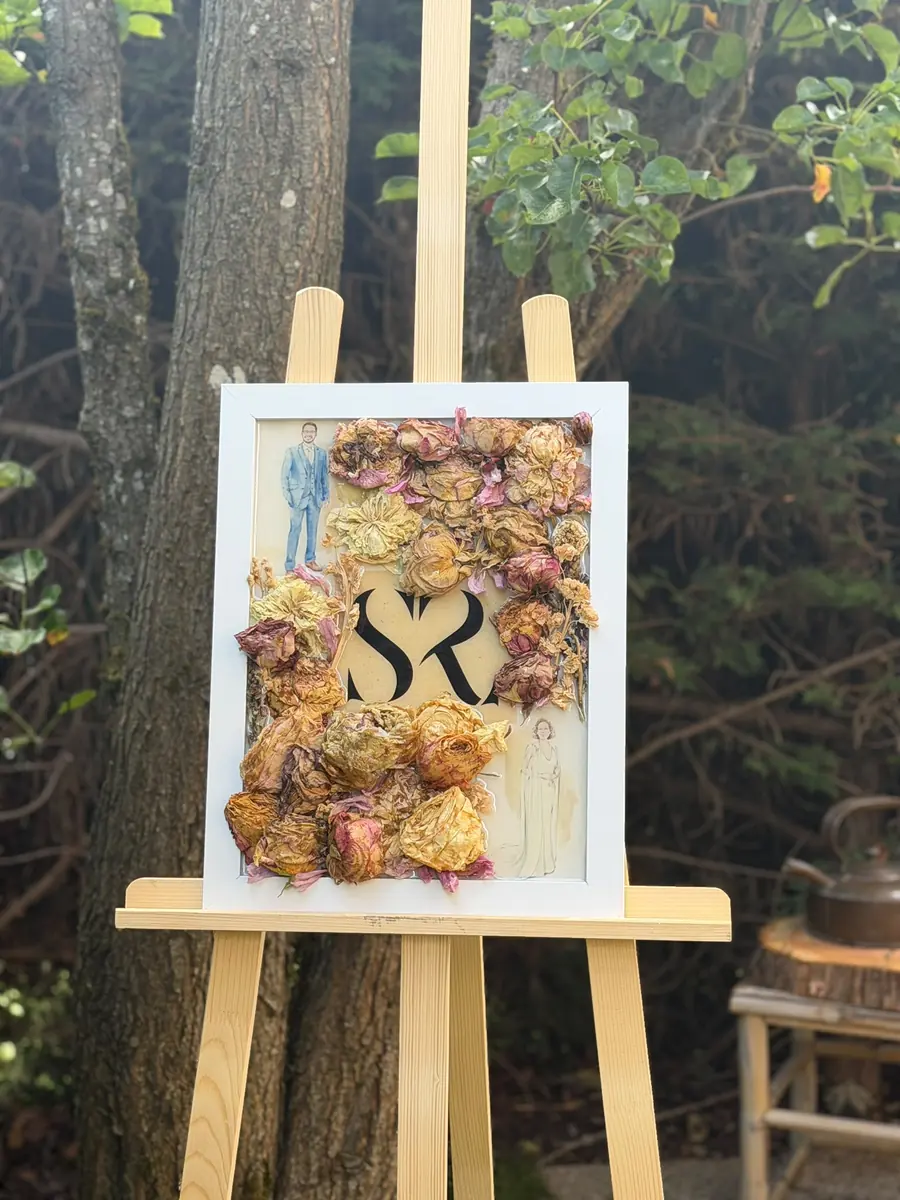 Cadre de mariage avec les roses du bouquet éternisées, monogramme des mariés et illustration du couple, posé sur un chevalet en extérieur