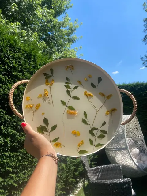 Main levant un plateau rond en résine avec des fleurs jaunes, contre un ciel bleu